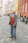 Per Arnoldi med stok står på brostensgade mellem farvede huse i byen