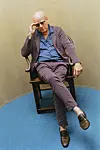 Thomas Blachman i stribet jakkesæt sidder i stol på blåt gulv og holder hånden for panden.