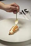 Hånd der hælder cremet sauce over en lille skaldyrsret på stor hvid tallerken